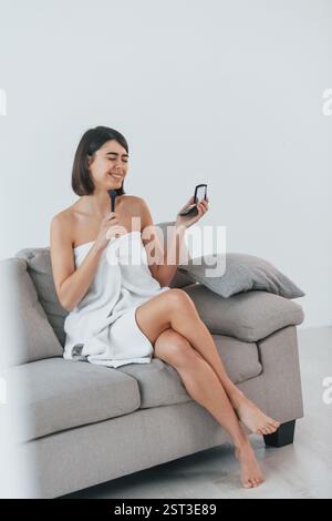 Positive female doing make up when sitting on the sofa. Young woman is at home. Conception of beauty. Stock Photo