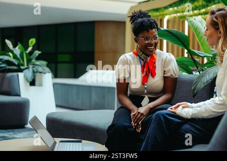 Two professional women engage in a friendly discussion while seated on a sofa in an office environment. The setting suggests a casual meeting amidst a Stock Photo