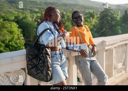 Laughing black friends chatting while lean on railing, waiting for taxi after booking on smartphone Stock Photo