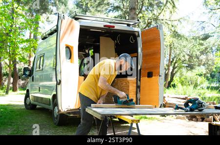 Carpenter using electric plane on wood plank, customizing camper van in forest Stock Photo