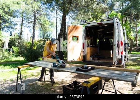 Carpenter using electric plane on wooden plank near open camper van, enjoying outdoor woodworking Stock Photo