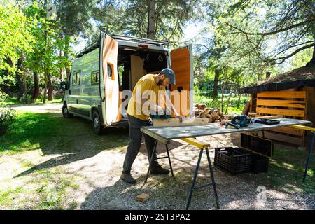 Carpenter using electric plane on wooden plank near his camper van in a forest, enjoying outdoor woodworking Stock Photo