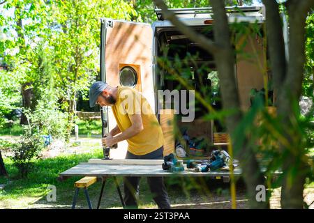 Carpenter working on wood plank near his camper van using a cordless drill, enjoying outdoor lifestyle Stock Photo