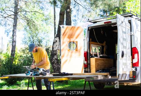 Carpenter working on a camper van conversion project outdoors, using power tools and surrounded by nature Stock Photo