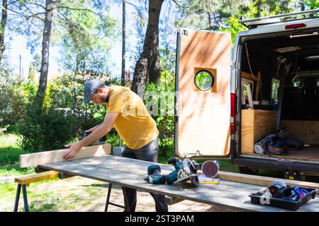 Carpenter working on wood plank using electric plane outdoors, near his van with open doors and tools inside Stock Photo