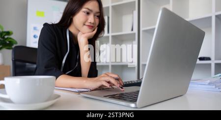 Focused woman analyzing financial data on a laptop while enjoying coffee in a modern office. Stock Photo