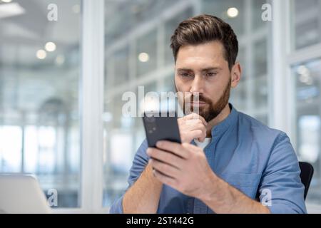 Serious man sitting with phone in hands, businessman thinking and reading online email. Company employee close-up at workplace inside office. Stock Photo