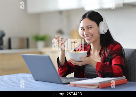 Happy asian student eating cereals while e-learning looking at you in the kitchen at home Stock Photo