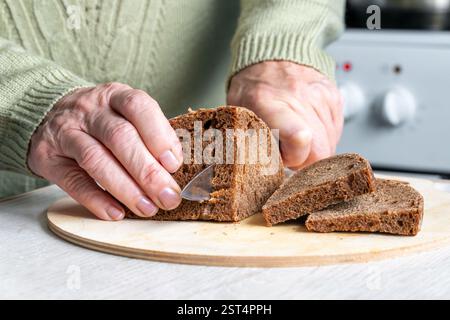 An elderly man is cutting rye black bread on a wooden chopping board in the kitchen. Stock Photo