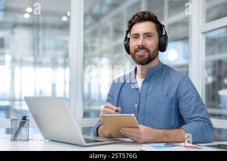 A man is seen watching a webinar and taking notes in a bright, modern office setting, with a laptop on the desk Stock Photo