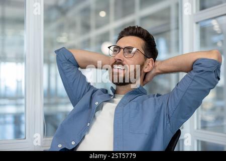 A thoughtful, bearded man relaxes with his hands behind his head, looking towards the window in a modern, bright office space Stock Photo
