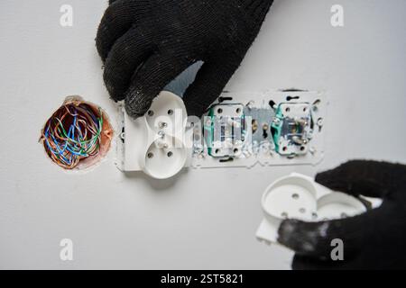 Close-up of an electrician wearing black gloves installing power socket on white wall. Electrical repair and maintenance process in progress Stock Photo