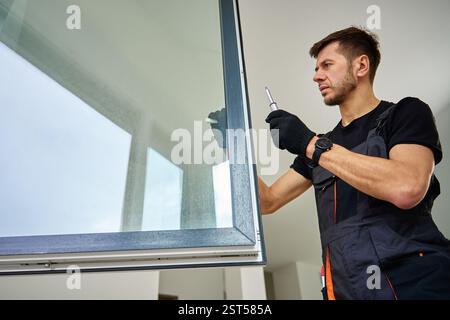Skilled worker wearing gloves uses screwdriver to fix window frame, home renovation tasks. Professional repairman adjusting white pvc windows in moder Stock Photo