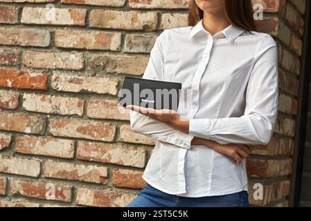Woman stylishly presenting an elegant black leather wallet in front of a brick wall, showcasing a fashion accessory, concept Stock Photo