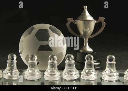 Small metal cup with ball of soccer or football sport made of glass on glass chess board with pawns as players Stock Photo