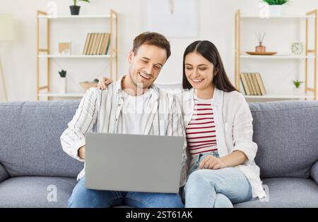 Happy young couple sitting on sofa in living room, hugging and using laptop for online entertainment Stock Photo