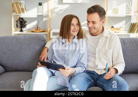 Happy young couple sitting on sofa in living room, smiling and using tablet and smartphone together. Stock Photo