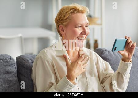 Happy mature woman holding smartphone making online video call at home. Stock Photo