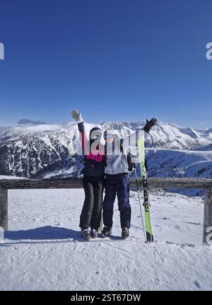 Happy couple standing on the snowy mountain top with arms raised posing as waving to camera. Skiers man and woman practice winter sports together havi Stock Photo