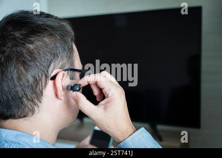 a man sticks headphones in his ear against the background of a TV screen.. watching TV with headphones at night. Stock Photo