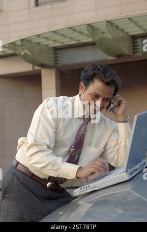 Young Indian Executive with tie, smiling and talking on mobile phone and working on laptop, on car in front of modern building Stock Photo