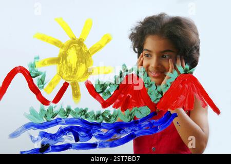 South Asian Indian eight year old girl child wearing red dress with shot hair painting with yellow, red, green, blue color on glass Stock Photo