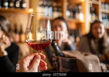 A mans hand holds a fine crystal glass of wine in the foreground with a wine cellar from Tuscany on a large unfocused wood shelves and other people. C Stock Photo