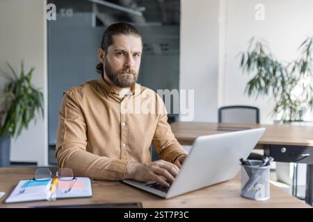 A bearded professional seated and typing on a laptop, displaying concentration and focus in a bright and modern office environment. Ideal for concepts related to business work, technology Stock Photo