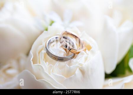 Two wedding rings are on top of a white flower. The rings are gold and silver. The flower is white and has green leaves Stock Photo