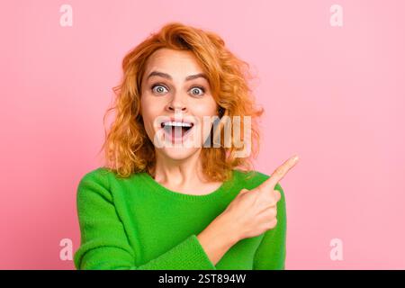 Excited young woman with red curly hair pointing aside against pink background wearing green jumper Stock Photo