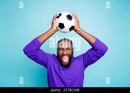 Photo of crazy excited mixed-race casual ecstatic black man raising soccer ball shouting with goal grimacing isolated vibrant blue color background Stock Photo