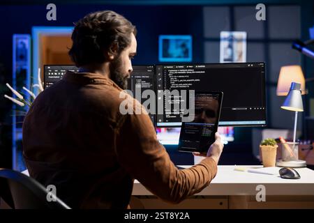 Programmer doing software debugging using tablet, checking glitches in lines of code displayed on computer monitors. Man working from home fixing coding issues using portable digital device Stock Photo
