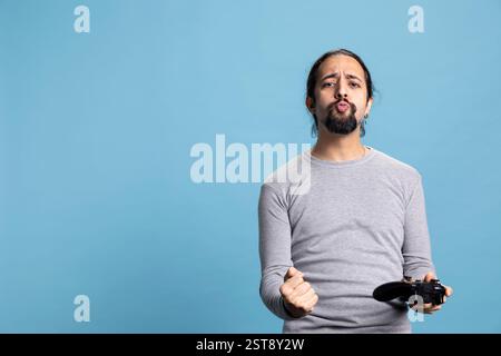 Overjoyed young adult winning an online tournament in the studio, feeling pleased and proud of his victory against blue background. Satisfied guy smiling in front of camera, video games on console. Stock Photo