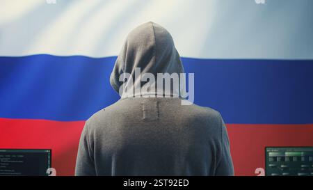 A Russian hacker working in a governmental hacking room with the Russian flag on big screen. Inflicting cyber terror, propaganda, fake news and misinformation in hybrid warfare. Camera B. Stock Photo