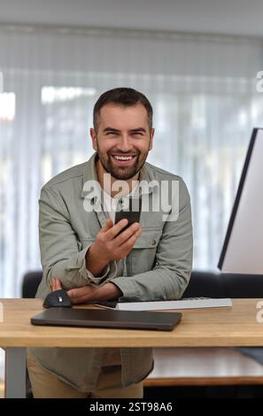 A young happy man using his phone mobile while working at home office, looking at the camera and smiling. Stock Photo
