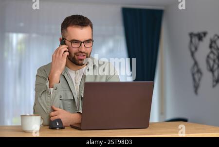 A happy man talking on his phone and using a desktop computer while working at home office. Stock Photo