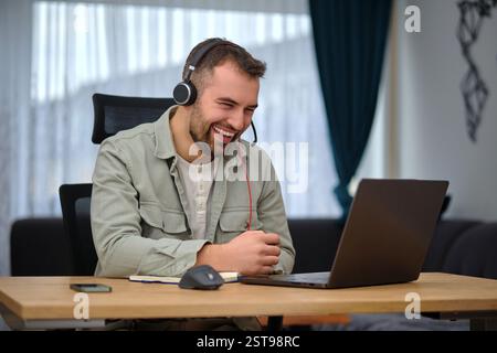 A happy man working at home office, wearing wireless headphones, having a video call with clients on a laptop, customer service or contact us support  Stock Photo