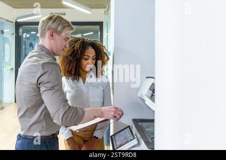 Two colleagues using a modern multifunction printer in a coworking office, enhancing productivity and teamwork Stock Photo
