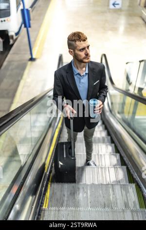 Businessman going up escalator in subway station, holding reusable coffee cup and pulling trolley suitcase Stock Photo