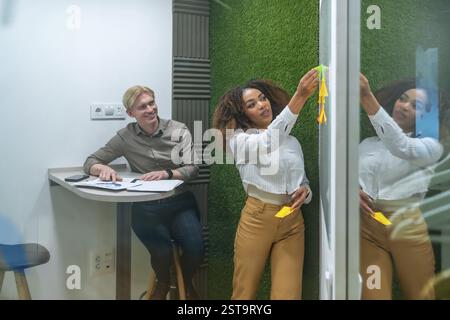 Businesswoman attaching sticky notes on a glass wall while brainstorming with a colleague in a modern coworking space Stock Photo
