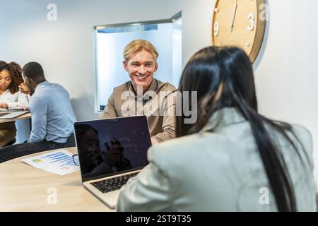Two managers are smiling and talking during a meeting in a coworking office, using a laptop and analyzing graphs Stock Photo