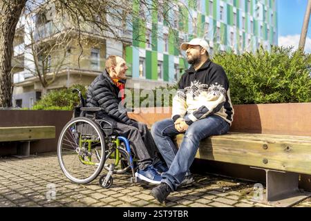 Two men, one in a wheelchair, enjoy a friendly conversation on a park bench in a modern urban setting Stock Photo