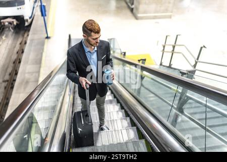 Young businessman going down an escalator in a station, carrying a suitcase and a coffee, going to work Stock Photo