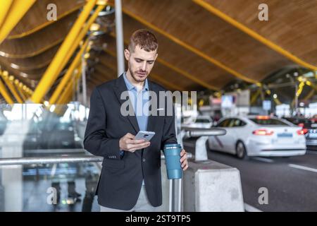 Young businessman using smartphone and holding reusable coffee cup while waiting for taxi or rideshare at airport Stock Photo