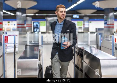 Young professional walking through subway turnstile holding reusable coffee cup, commuting to work in the morning Stock Photo