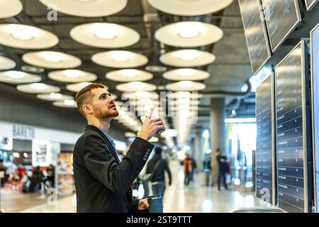 Businessman consulting his smartphone and checking flight information displayed on screens at the airport Stock Photo