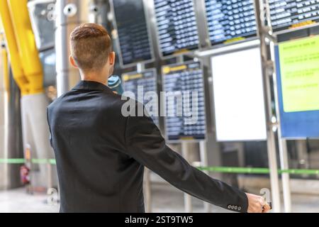 Businessman holding passport and pulling suitcase while checking flight schedule on departure board at airport terminal Stock Photo