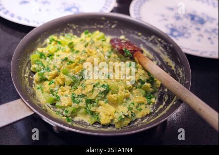 A skillet and wooden spoon with scrambled eggs with fresh greens and plates in the background on a stovetop. Stock Photo
