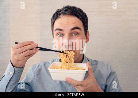 A young, handsome, cheerful man is eating delicious instant noodles with chopsticks Stock Photo