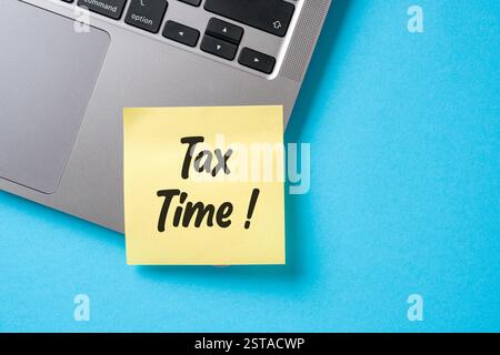 Tax Time written on a sticky note stuck to the corner of a laptop on a blue desk Stock Photo